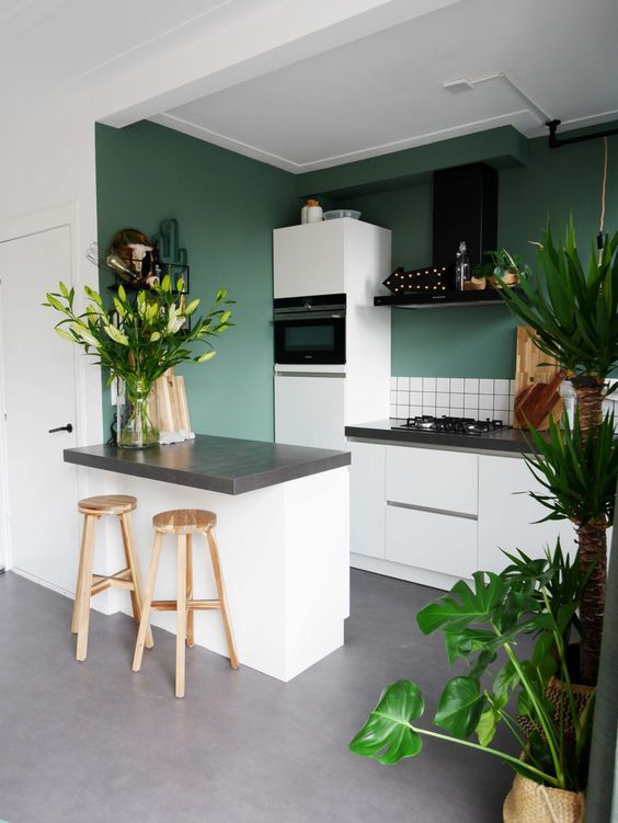 a green kitchen with white tiles, gray stone countertops, a black extractor hood and potted plants and flowers