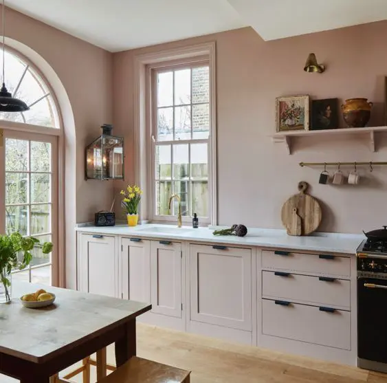 A beautiful dusty pink kitchen with matching cabinets, shaker cabinets, black handles, white stone countertops and shelving