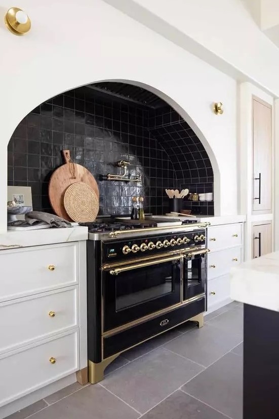 a chic vintage kitchen with white shaker cabinets, black zellige tiles, a black vintage stove and gold fixtures