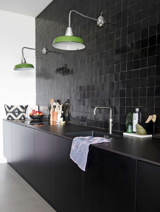 a cool black kitchen with sleek base cabinets, tiles all over the wall, green lamps and stone countertops