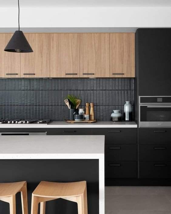 a minimalist black kitchen with stained upper cabinets, a black narrow tile backsplash, a white kitchen island and a black pendant light