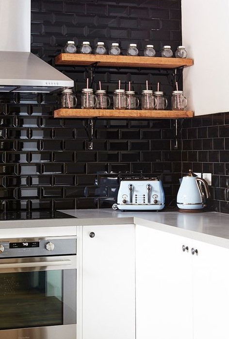 a white kitchen with gray countertops and a black glossy tile backsplash that takes up two walls, as well as floating, stained shelves for spices