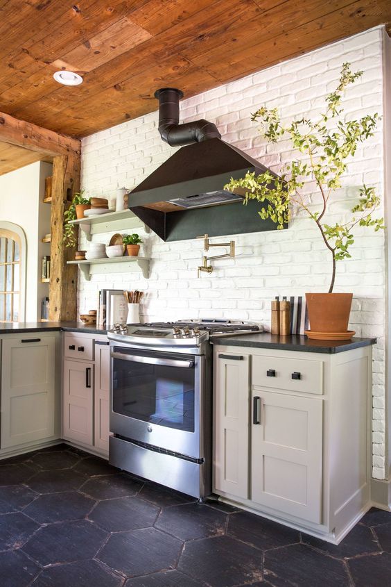 A dove gray kitchen with shaker-style cabinets, black countertops, a white brick wall and potted plants, and a black range hood