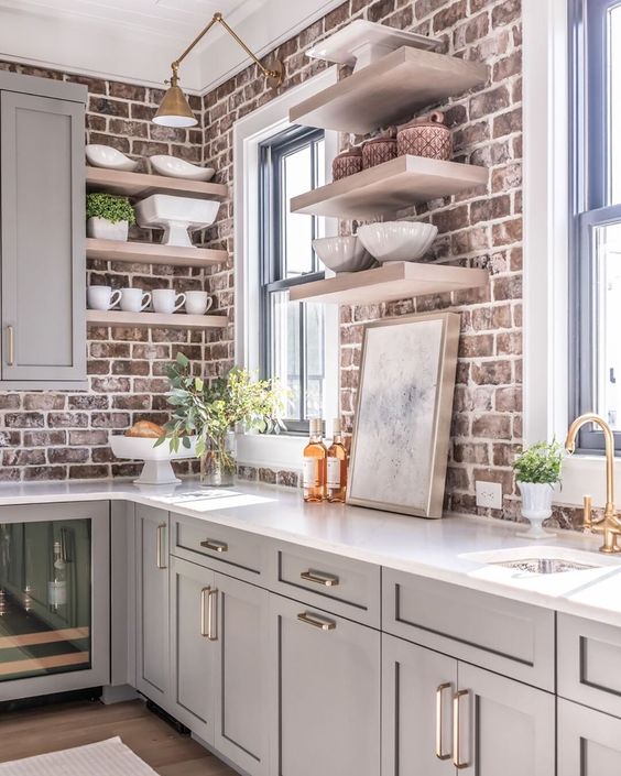 A dove gray kitchen with shaker-style cabinets, white countertops, open shelving, red brick backsplash, and brass accents