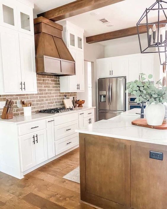 An inviting white farmhouse kitchen with shaker-style cabinets, a metal hood, dark-stained wood beams and a red brick backsplash