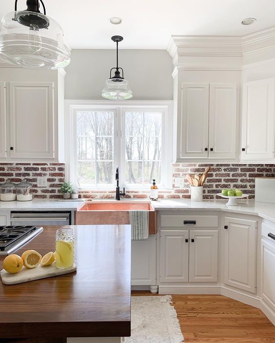 a white farmhouse kitchen with shaker-style cabinets, a red brick backsplash, white stone countertops and an island with a stained countertop