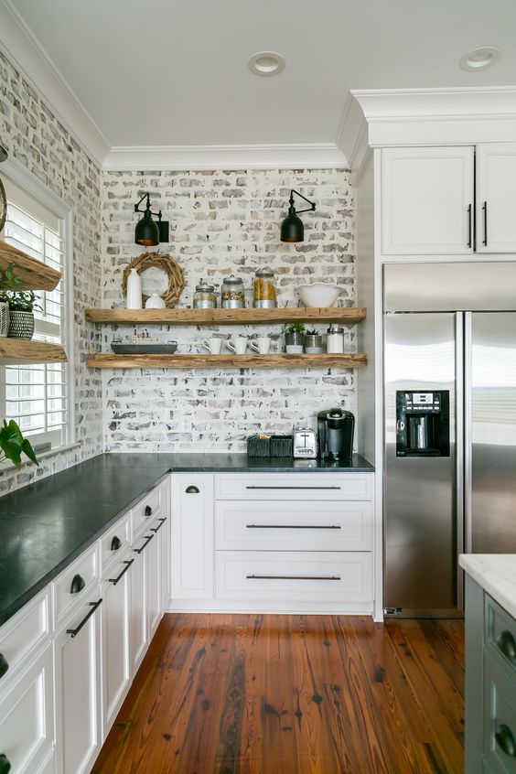 a white farmhouse kitchen with shaker cabinets, black stone countertops, white shelves and a whitewashed red brick backsplash