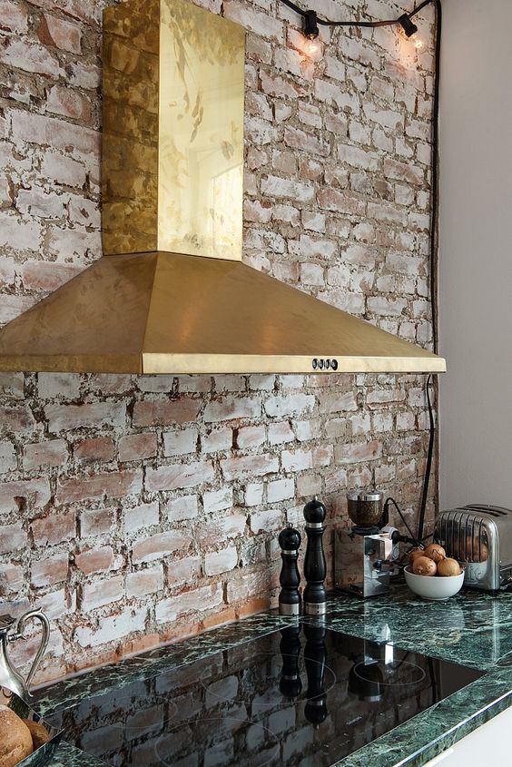 A back wall made of whitewashed brick harmonizes with a shiny gold hood and a green stone countertop, giving the room a historical feel