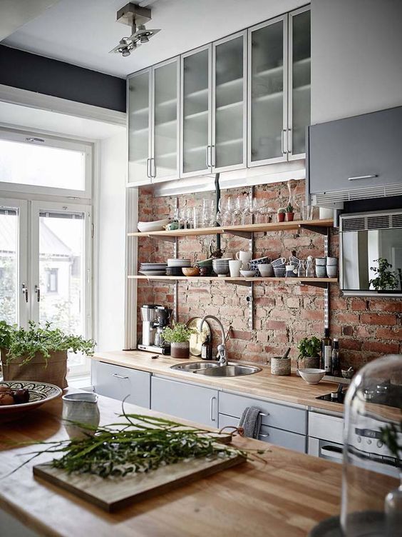 A sleek gray Scandinavian kitchen with sleek butcher block countertops, a rough red brick backsplash, frosted glass cabinets and lots of natural light