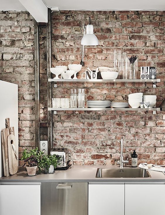 A clean white Scandinavian kitchen with sleek cabinets, concrete countertops and a rough red brick backsplash that adds interest and chic to the room