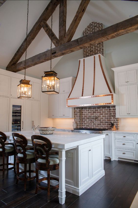 a white vintage kitchen with shaker style cabinets, built-in lights and a large range hood with copper details, a red brick backsplash for a touch of color and interest, pendant lamps and dark stained stools