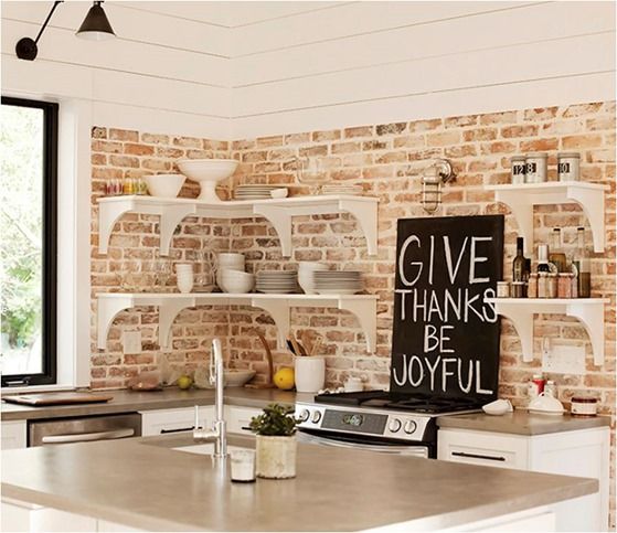 a white kitchen with shaker-style cabinets and black handles, with open shelving, a whitewashed brick backsplash, and a small kitchen island with a concrete countertop