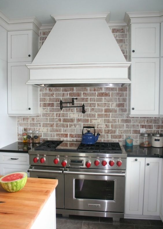 a white kitchen with shaker-style cabinets, a large range hood and a whitewashed red brick backsplash that adds interest, texture and color to the room