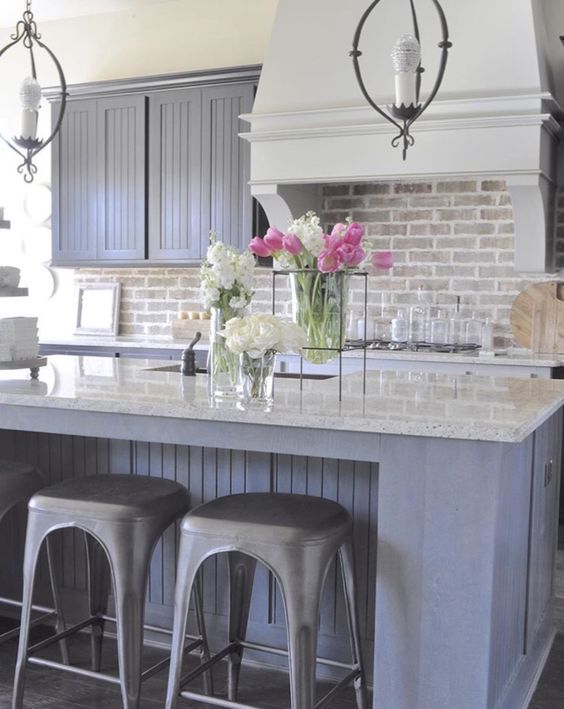 a farmhouse kitchen with gray beadboard cabinets, a brick backsplash and a white range hood, white countertops and vintage pendant lamps
