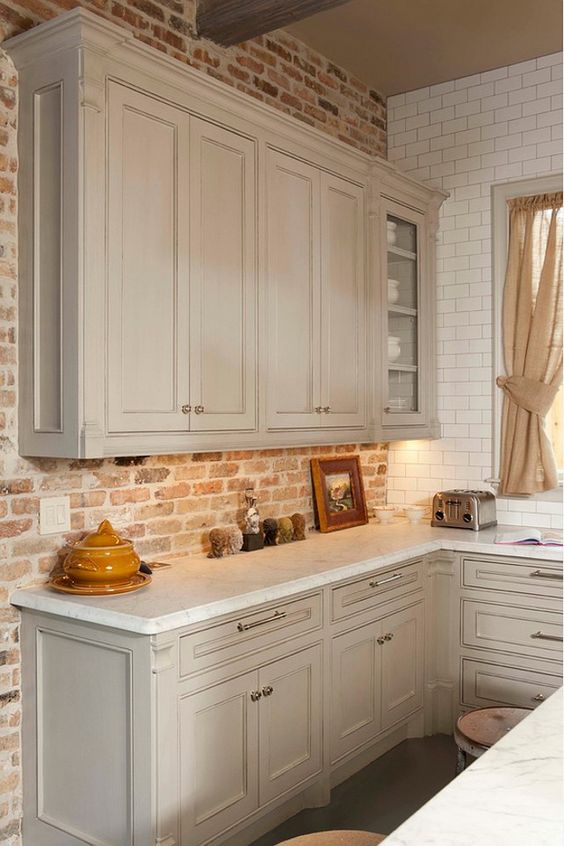 A sleek dove gray kitchen with shaker-style cabinets, white countertops, a red brick backsplash, and neutral curtains is a beautiful idea for an eclectic home