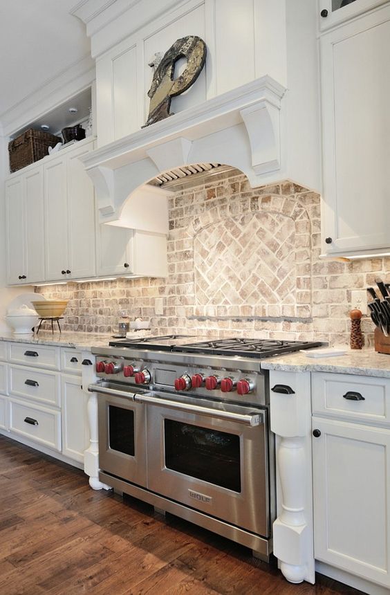 A white farmhouse kitchen with shaker-style cabinets, black knobs, a whitewashed patterned brick backsplash, and built-in light fixtures and stainless steel appliances