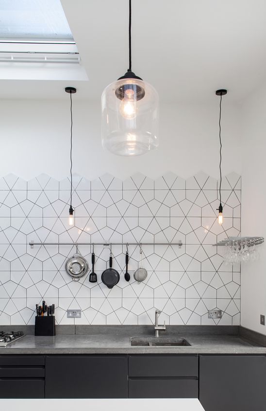 a minimalist black kitchen with matte cabinets, gray countertops, a black-and-white geometric tile backsplash, and pendant lamps