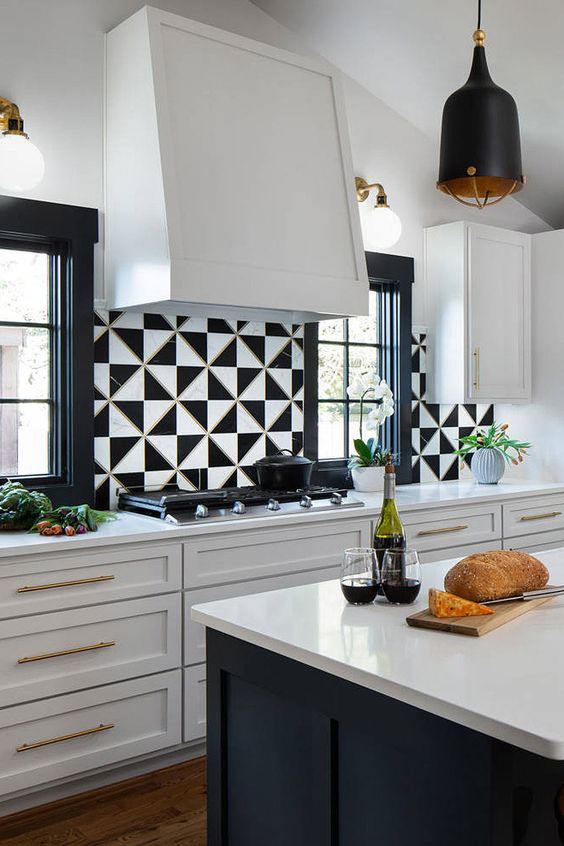 a sleek white kitchen with shaker style cabinets, a black kitchen island with white stone countertops, black and white geotiles and a large hood
