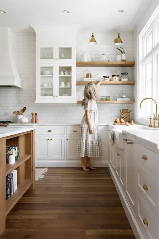 A cozy, modern kitchen with white cabinets, light-stained shelves, a light-stained kitchen island, wall sconces, and brass fixtures is chic