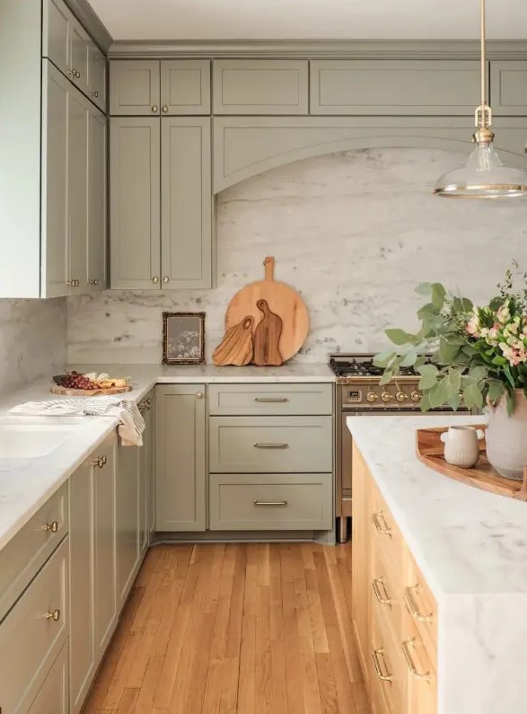 A beautiful sage green kitchen with shaker cabinets, a white stone backsplash and countertops, a stained kitchen island, and a chic pendant lamp