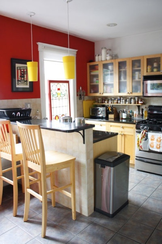 a small, bright kitchen with a striking red wall, yellow pendant lamps, light stained furniture and a high countertop for dining