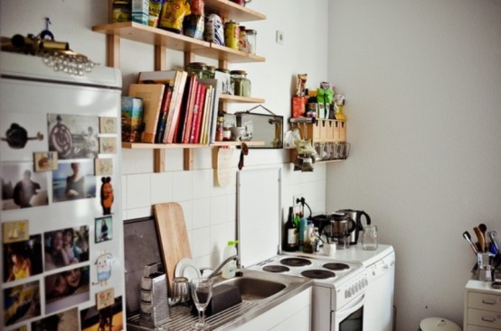 a small modern kitchen in white with light stained wooden shelves and lots of personal photos