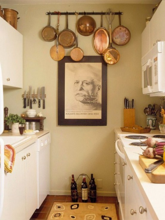 a small neutral kitchen with cream cabinets, metal knobs, a piece of art and a rack of vintage pans