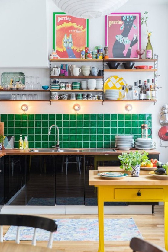 a maximalist kitchen with black sleek cabinets, an emerald green tile backsplash, a yellow table, colorful artwork and open shelving
