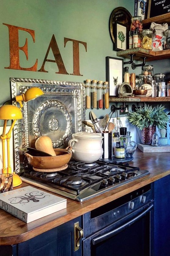 a maximalist kitchen with green walls and navy blue cabinets, butcher block countertops, rough wood shelves and a yellow lamp