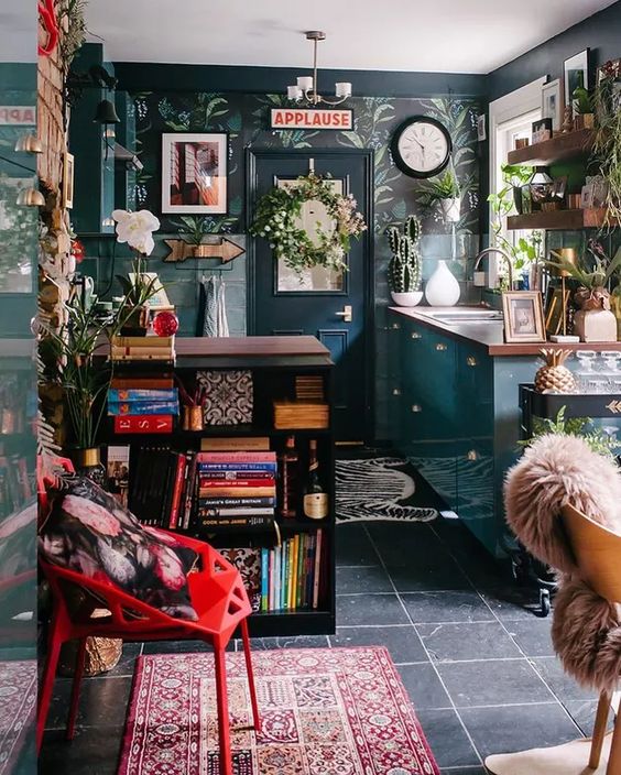 an eclectic and maximalist kitchen with dark wallpaper, teal cabinets, a black bookcase, some shelves and green plants, and a statement rug