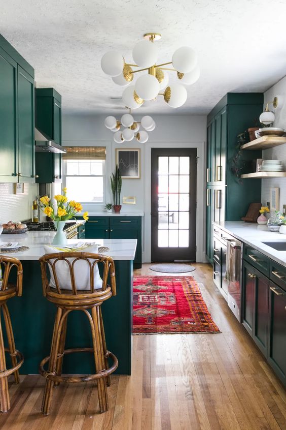 An eclectic kitchen features emerald green cabinets, white marble countertops, tall rattan stools, modern chandeliers and a statement boho rug