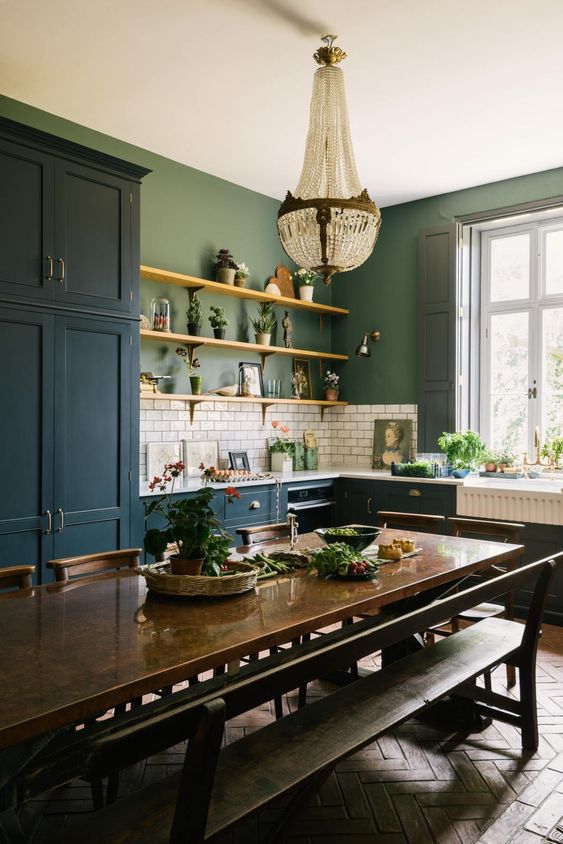 an eclectic kitchen with navy blue cabinets, green walls, a white subway tile backsplash, a dark stained table and benches, and a crystal chandelier