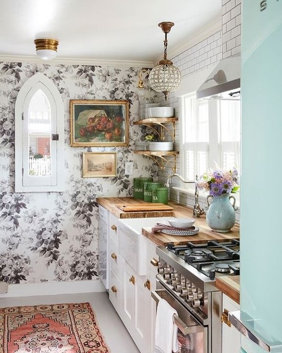 Traditional white cabinets with butcher block countertops are paired with a floral wall, glam lamp, and boho rug