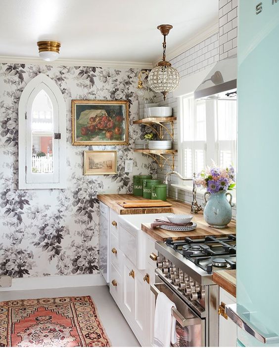 Traditional white cabinets with butcher block countertops are paired with a floral wall, glam lamp, and boho rug