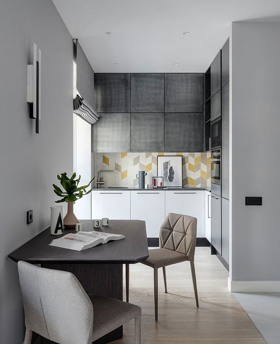 a small modern kitchen in gray and white with a light tiled splashback, built-in appliances and a dining area next to it