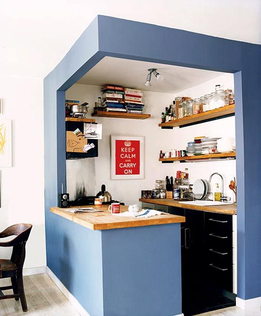 A small kitchen cube with blue walls, black cabinets, open shelves, and butcher block countertops is super cool