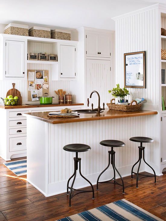 A small white farmhouse kitchen with stained wooden worktops, black stools, boxes and baskets looks inviting