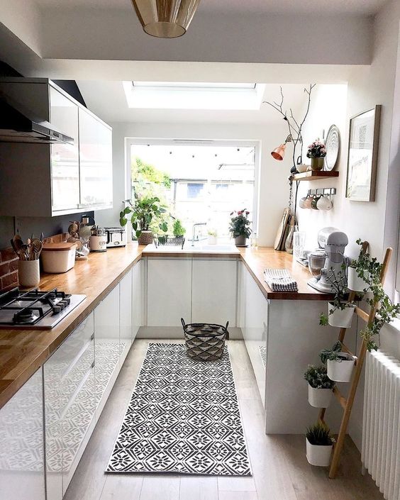 a small white kitchen with butcher block countertops, potted plants, a printed rug, and pendant lamps