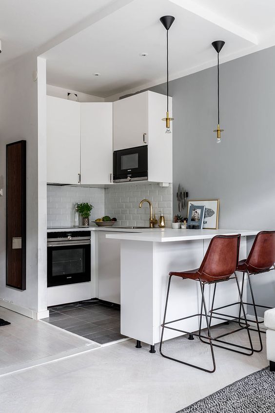 A small white kitchen with white tiles, pendant lamps, brown leather stools and gold accents is chic