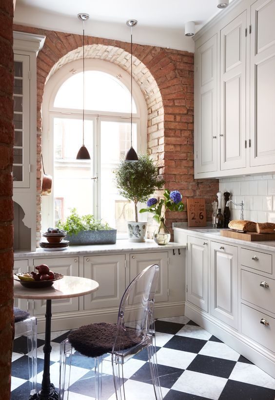 a white farmhouse kitchen with a windowsill and potted plants, a round table, ghost chairs and a white tile backsplash