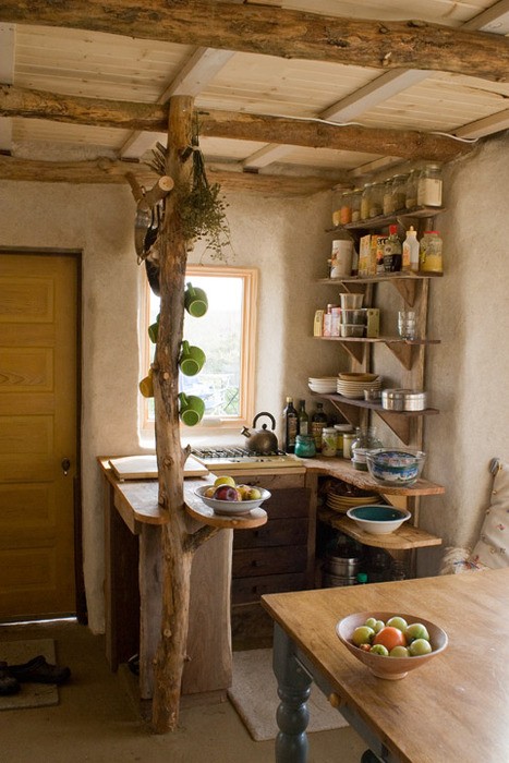 a small wabi-sabi kitchen with open shelves, rough wooden beams on the ceiling and a column with cups, an additional table that can be used as a kitchen island
