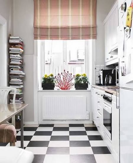 A small white kitchen with checkered floors, butcher block countertops, a bright striped curtain, and a stand of books is cozy