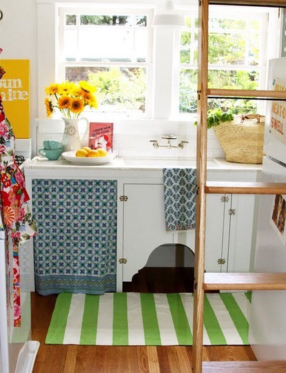 a tiny cottage kitchen with white cabinets, printed colorful textiles and a ladder for storing things
