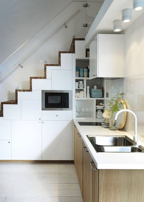 a Scandinavian kitchen with lower wooden cabinets, a staircase with white storage compartments, a glossy tile backsplash and spotlights