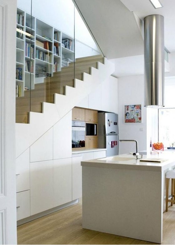 A small modern kitchen in white, built under the stairs, with a large kitchen island, an extractor hood above and some appliances