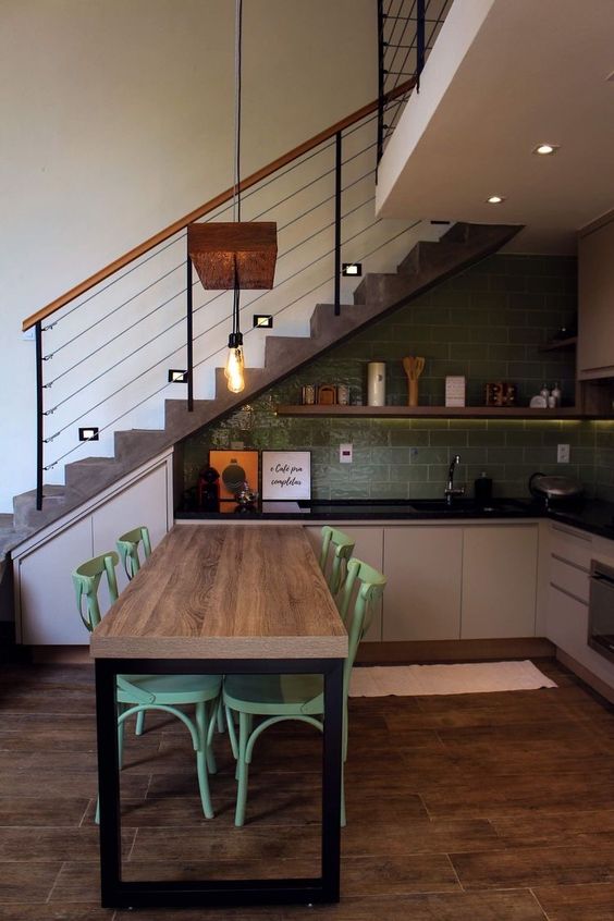 a small industrial kitchen with gray cabinets, black countertops, a green subway tile backsplash, a dining area and pendant lamps