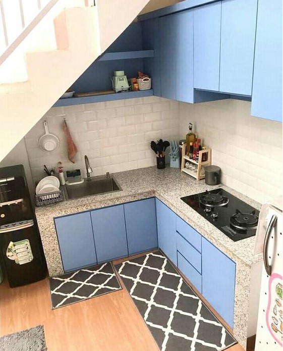 a tiny blue kitchen under the stairs with a subway tile backsplash, terrazzo countertops and black rugs