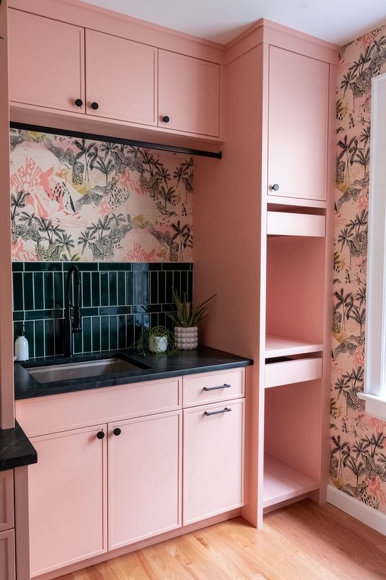 a bright pink kitchen with cabinets and open compartments, black countertops, dark green tiles and floral wallpaper