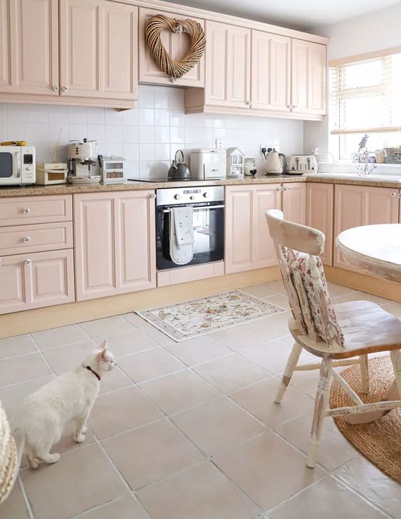 a pretty blush kitchen with shaker cabinets, stone countertops, a white square tile backsplash, a round table and shabby chic chairs