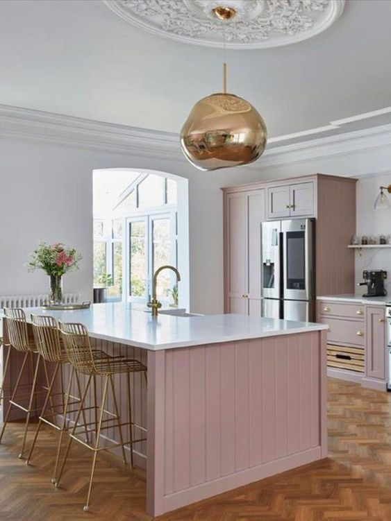 a pale pink kitchen with overlapping cabinets and white stone countertops, as well as a white stone backsplash, a gold pendant lamp and matching high stools
